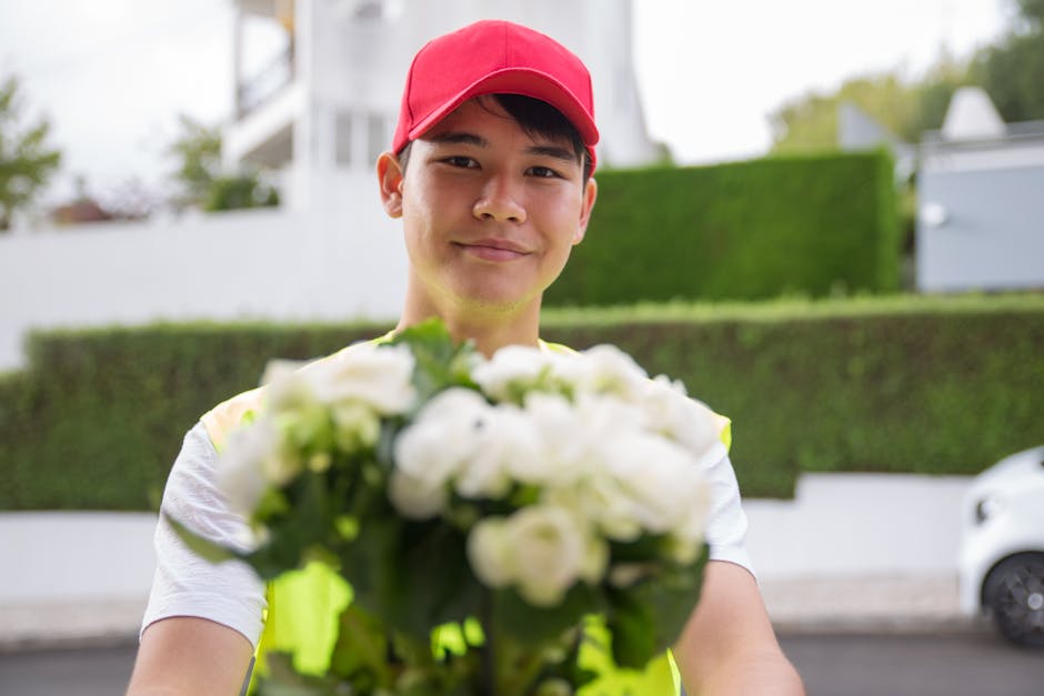 A smiling delivery person wearing a red cap and a high-visibility vest holds out a bouquet of fresh white roses and green foliage towards the camera. The background features a suburban street with nea