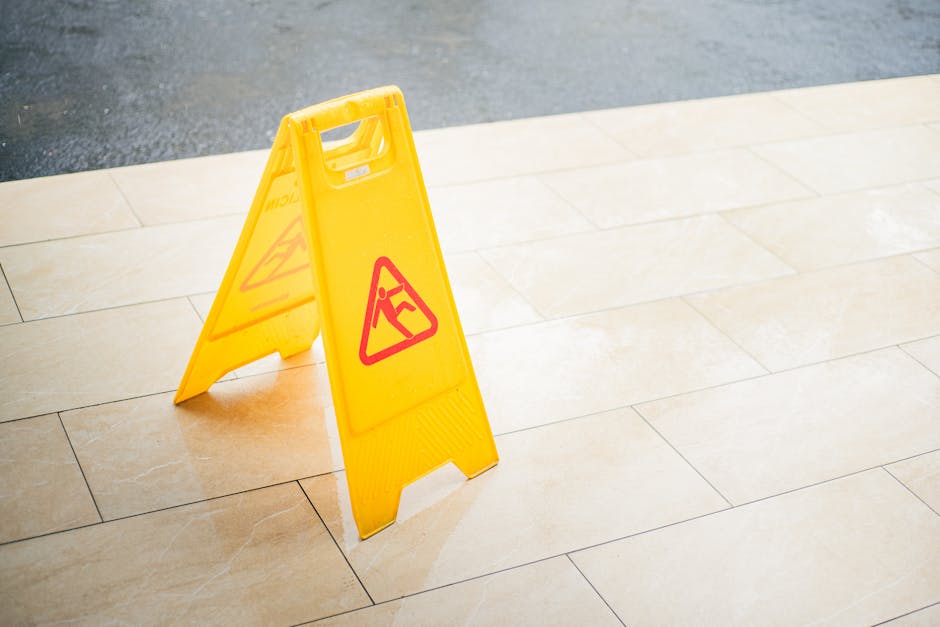 A yellow portable wet floor warning sign with red and black symbols standing on a beige tiled floor indoors. The sign indicates a caution for a slippery surface and is positioned near the edge of the
