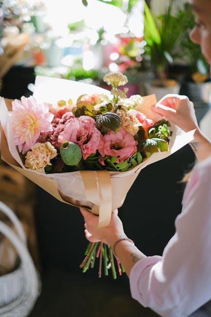 A person holding a large bouquet of fresh flowers, including pink peonies, white carnations, and purple chrysanthemums, wrapped in cream and white paper with a satin ribbon. The bouquet features lush
