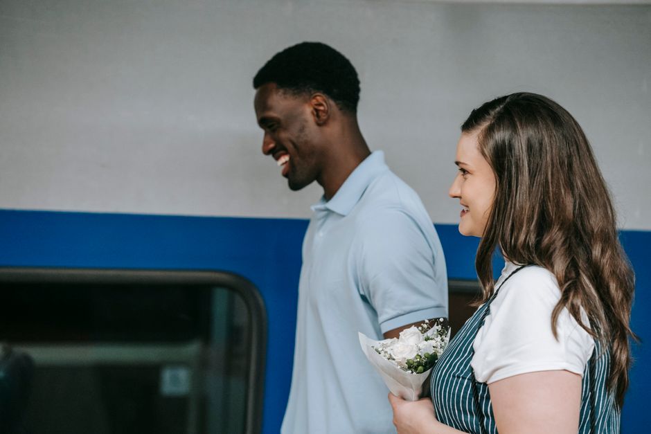 A smiling woman with long brown hair in a white blouse and striped apron is holding a small floral bouquet wrapped in clear cellophane, featuring white and green flowers. Standing beside her is a man 