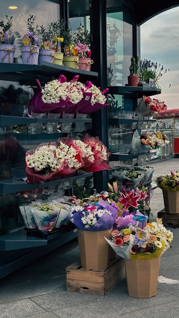 A display of fresh flower arrangements outside a florist shop, featuring various bouquets with white, pink, purple, and yellow blooms. Some flowers are wrapped in colorful tissue paper and cellophane,